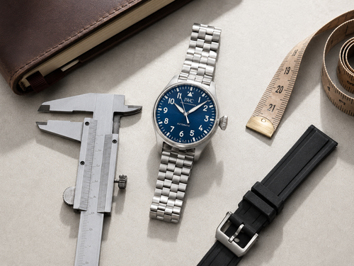 Overhead technical view of an IWC Big Pilot watch with blue dial on a steel bracelet alongside calipers, measuring tape, and a black EasX-CHANGE strap